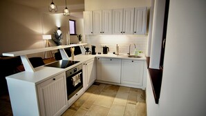 Eine moderne Küche mit weißen Schränken, einer schwarzen Arbeitsplatte und einem eingebauten Ofen. Es gibt eine Spüle mit Armatur, eine Kaffeemaschine und einen Wasserkocher. Der Boden ist mit Fliesen ausgelegt, und über der Arbeitsplatte befinden sich zwei Pendelleuchten.