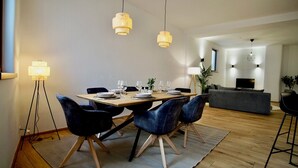 Ein Essbereich mit einem Holztisch, gedeckt für vier Personen, umgeben von dunkelblauen Stühlen mit Holzbeinen. Über dem Tisch hängen zwei Pendelleuchten.