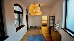 Ein Korridor mit Holzboden, einem farbenfrohen Teppich, einer Korbschwingerleuchte, einem schwarzen Metallregal und einem Fenster mit Blick auf Bäume.