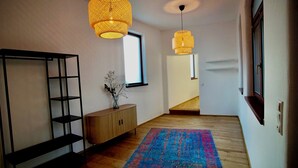 Ein Flur mit Holzboden, einem blau-roten Teppich, einer schwarzen Regaleinheit, einem Holzkonsole mit einer Vase weißer Blumen und zwei Pendelleuchten.