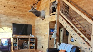 Interior de cabana de madeira com uma televisão grande, móveis de couro, cabeça de veado montada e uma escada.