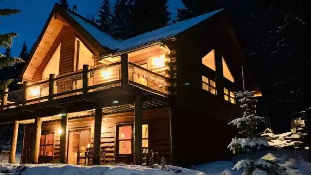 Uma cabana de toras com varanda e janelas amplas, rodeada por neve.