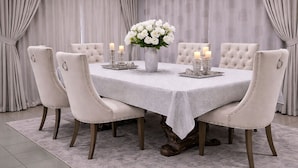 Un comedor con una mesa redonda, seis sillas tapizadas, un jarrón de flores blancas y candelabros.