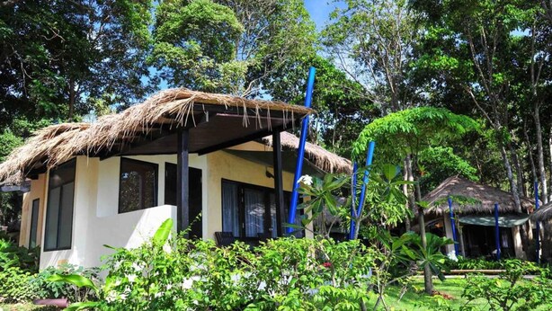 A thatched-roof structure with a white exterior, surrounded by lush greenery and trees.