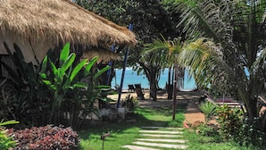 Un giardino tropicale con una struttura a tetto di paglia, palme e un sentiero in pietra che conduce a una spiaggia.