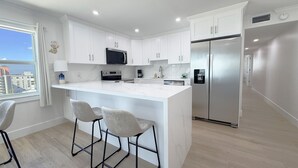 A modern kitchen with white cabinets, a stainless steel refrigerator, a microwave, and a large island with bar stools. - Gateway Villas #899 "Sunset Beach Penthouse" (Fort Myers Beach)