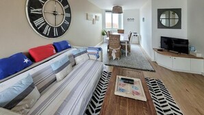 A living room with a sofa, a coffee table, a TV, and a large wall clock. - Superbe appartement vue mer (Dunkerque)