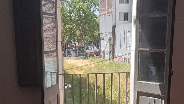 Een kamer met een open schuifglazen deur die uitkomt op een balkon. Het balkon heeft een metalen hekwerk en uitzicht op gebouwen en bomen.