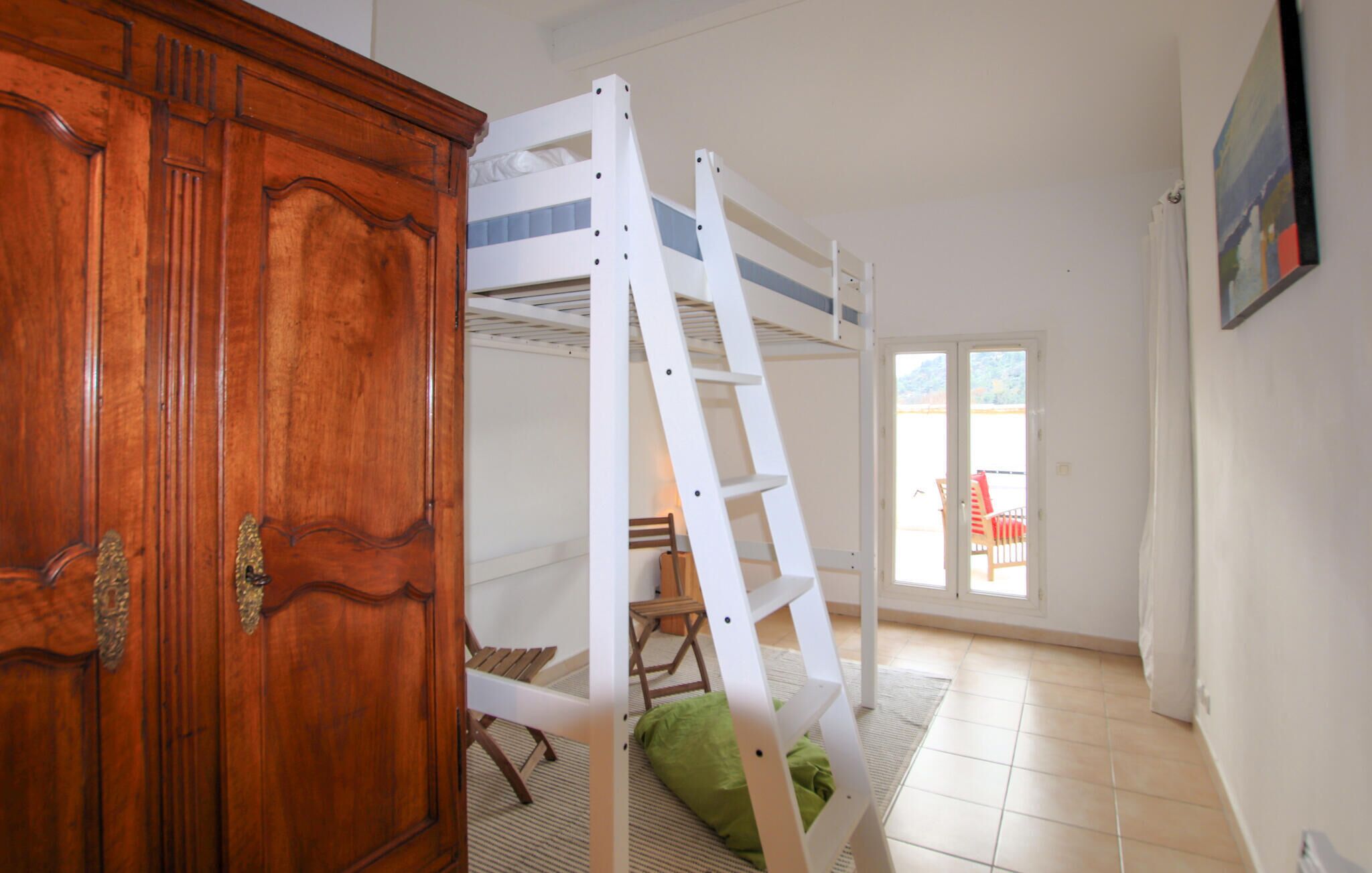 Una habitación con literas, un armario de madera, un cojín verde y vistas a un balcón con una silla.