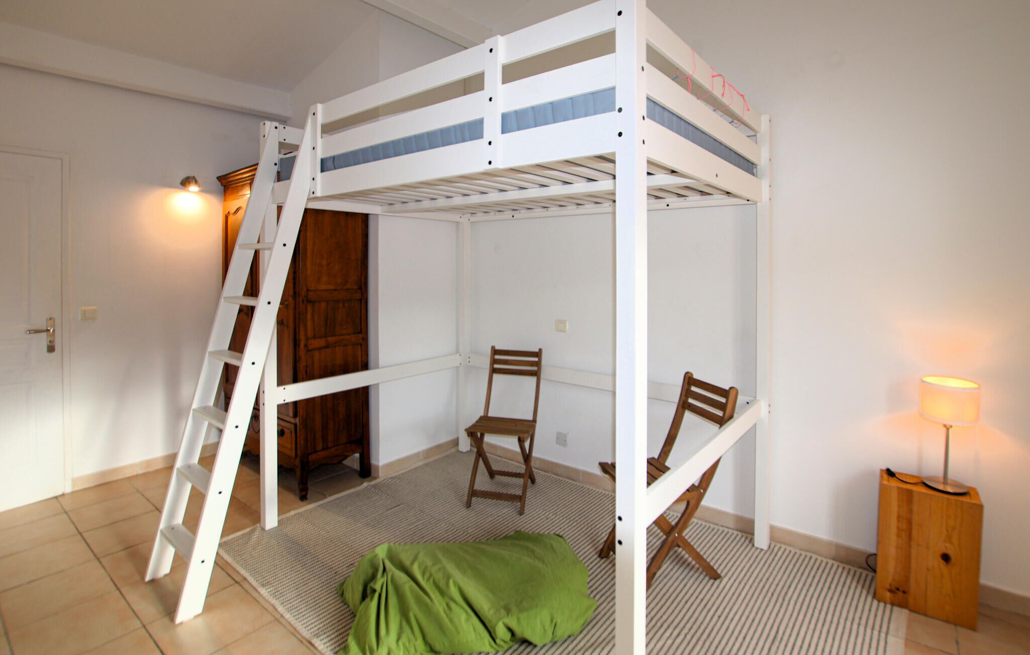 A bunk bed room with a ladder, two wooden chairs, a green blanket, and a lamp.
