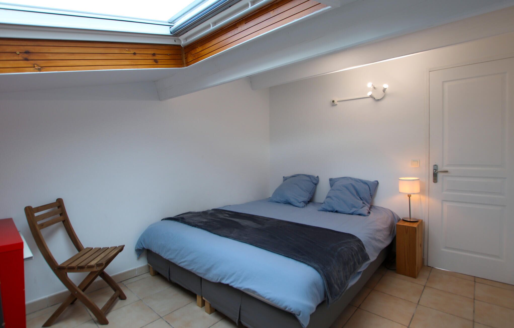 Une chambre avec un puits de lumière, un lit avec une literie bleue, une chaise en bois et une table de chevet avec une lampe.