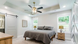Quarto com cama, mesa de cabeceira, cômoda, ventilador de teto e janela com vista para o verde.