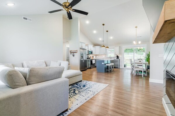 A modern living room with a sofa, a ceiling fan, and a kitchen in the background. - TV Per Room, 3bd Centrally Located Near Ft Riley (Manhattan)
