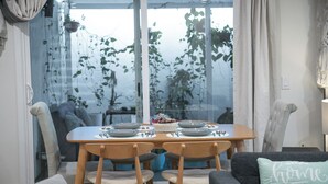 Een eetgedeelte met een tafel voor vier personen, stoelen, een raam met planten en een kussen met tekst.