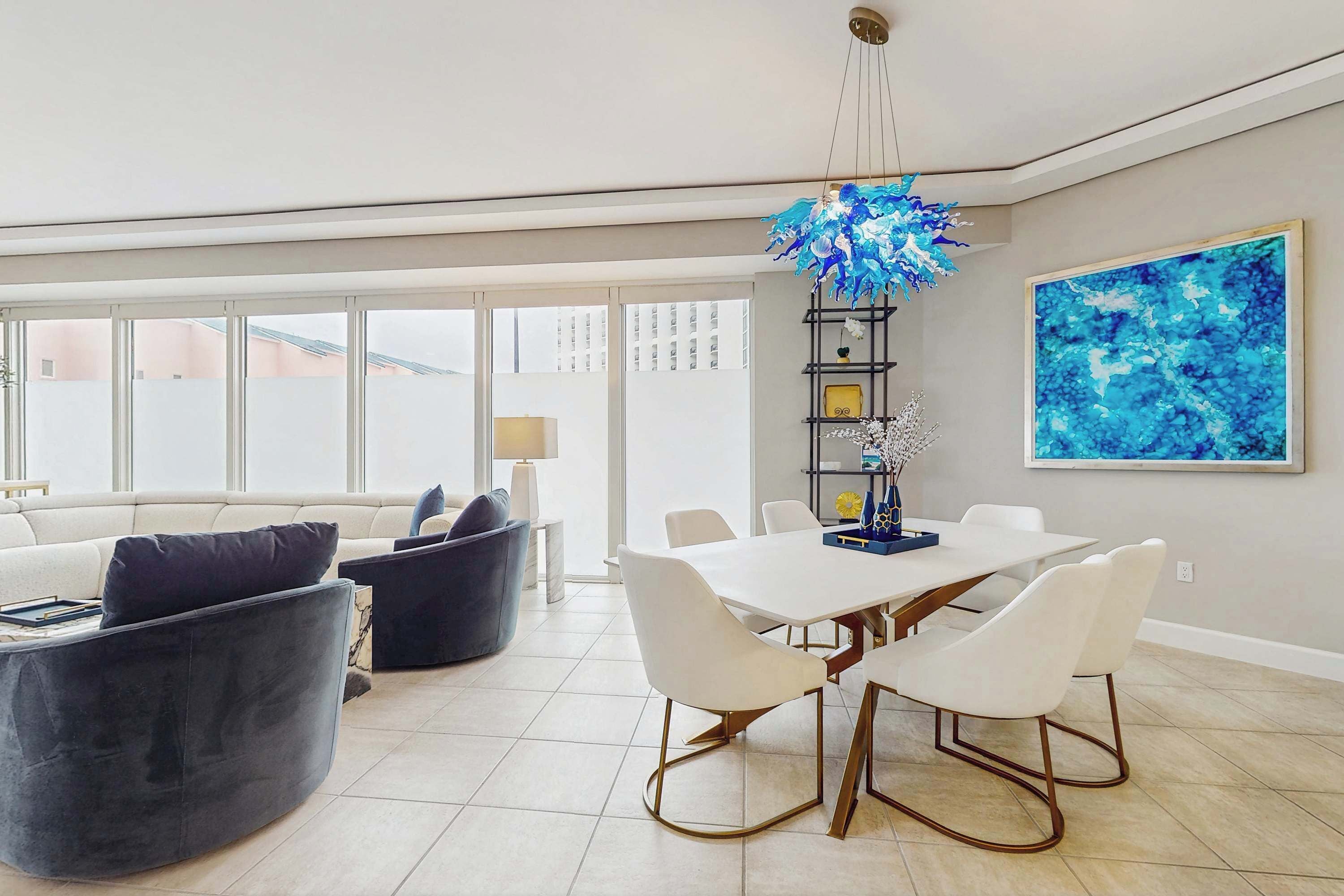 A modern living room with a dining area, featuring a white dining table, white chairs, and a blue abstract painting on the wall.
