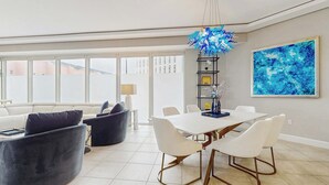 A modern living room with a dining area, featuring a white dining table, white chairs, and a blue abstract painting on the wall. - Exclusive Gulf view 3BR, pool & tennis (Miramar Beach)