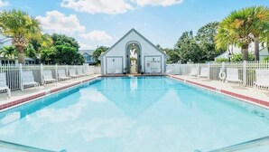 Ein rechteckiger Swimmingpool, umgeben von einem weißen Zaun, mit einem kleinen Gebäude mit Bogenöffnung und Palmen im Hintergrund.