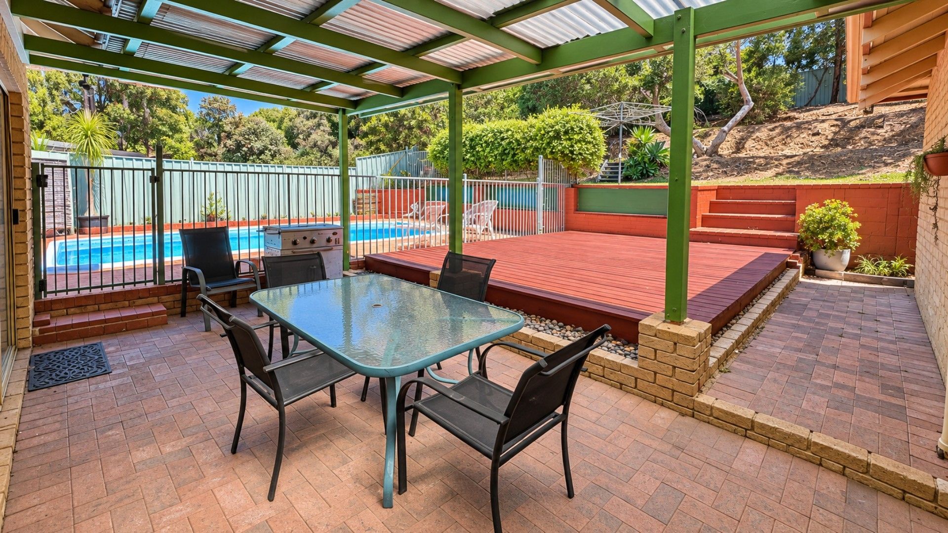 A covered patio area with a glass table and chairs, a barbecue grill, and a swimming pool in the background.