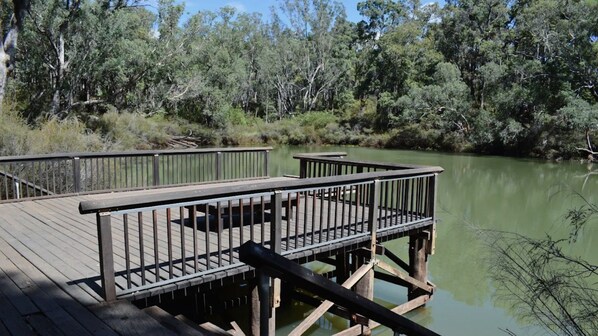 Une terrasse en bois, dotée de rampes qui s’étendent au-dessus d’une étendue d’eau, et entourée d’une végétation luxuriante.