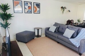Ein Wohnzimmer mit grauem Sofa, einem kleinen Hocker, einem runden Beistelltisch und drei abstrakten Bildern an der Wand.