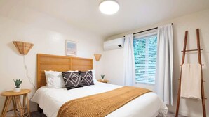 Ein Schlafzimmer mit einem großen Bett, einem hölzernen Kopfteil, einem Fenster mit Jalousien, einer stehenden Leiter und einem kleinen runden Tisch mit einer Pflanze.