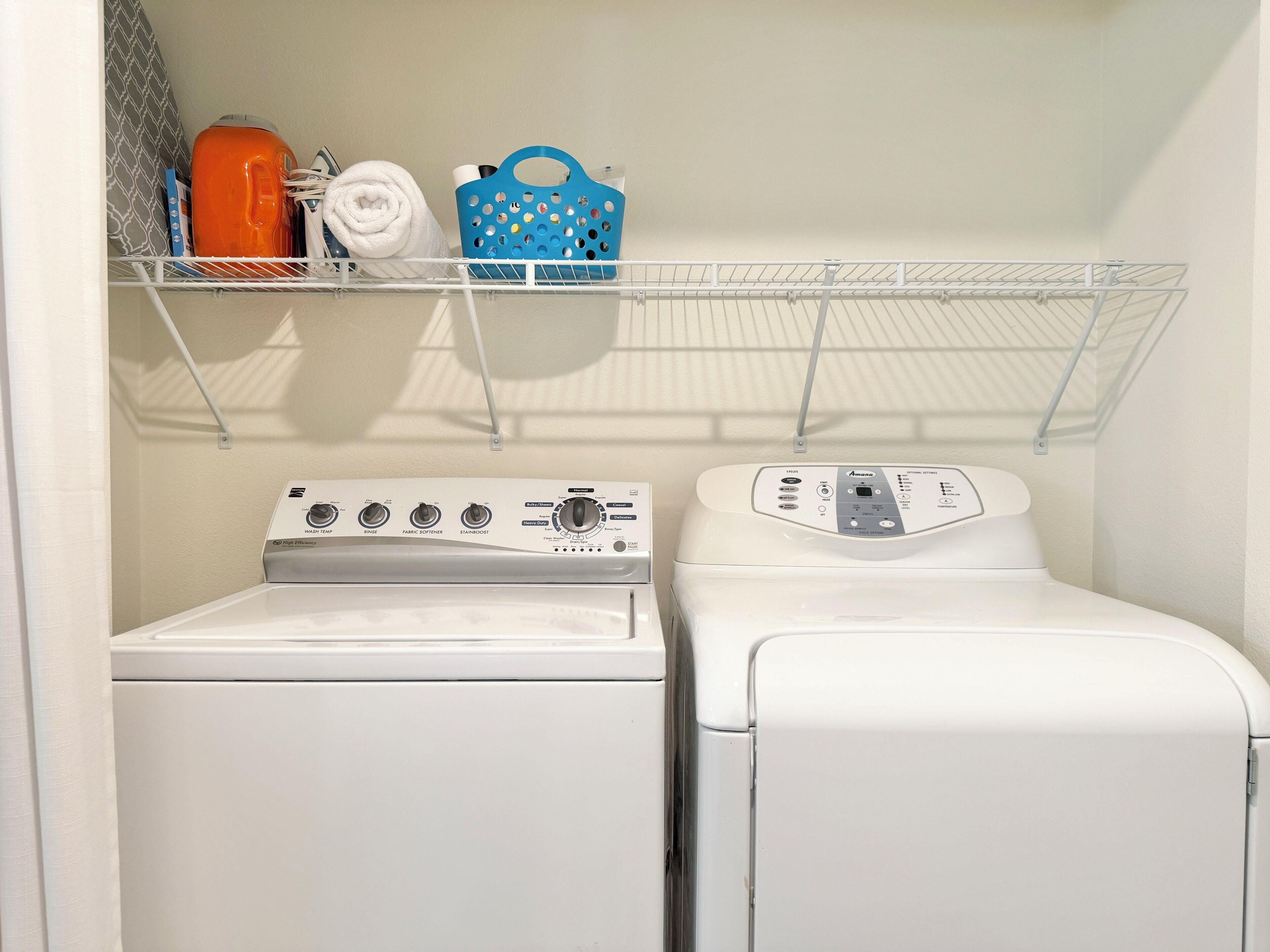 A laundry area with a front-loading washer and dryer, a shelf with a blue basket, and a rolled-up towel.