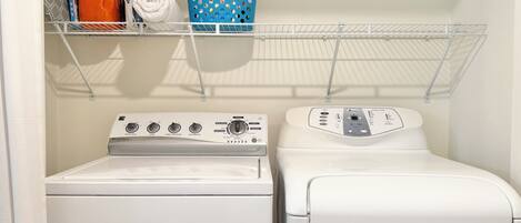 A laundry area with a front-loading washer and dryer, a shelf with a blue basket, and a rolled-up towel.