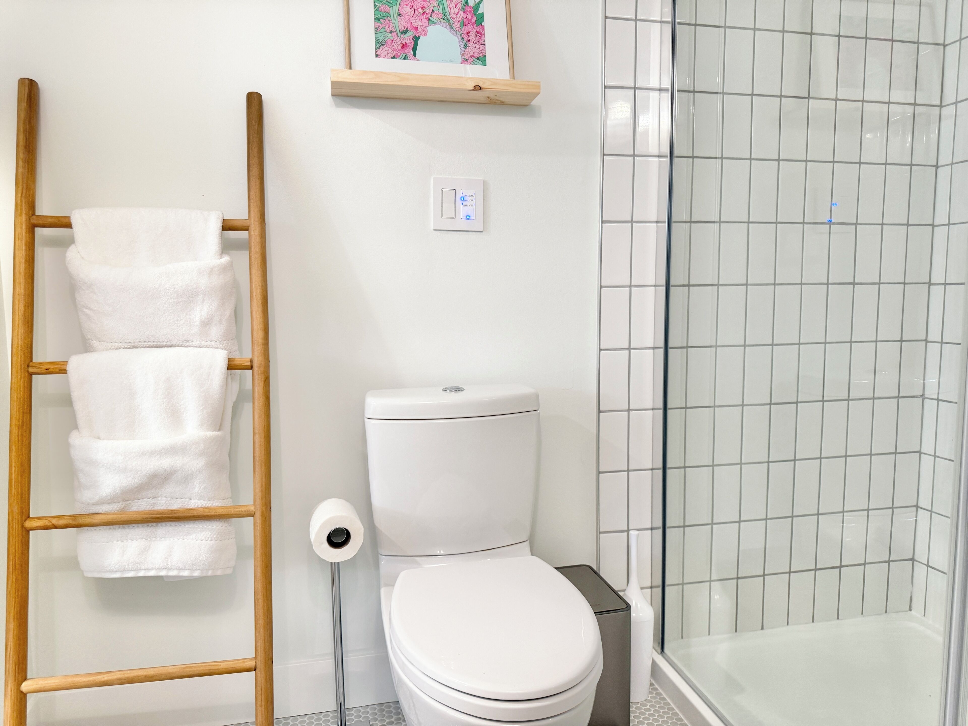 A bathroom with a toilet, a wooden towel rack with towels, a toilet paper holder, a picture frame, and a tiled shower area.