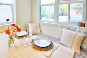 A dining area with a table set for two, a vase of flowers, and a view of the backyard through large windows.