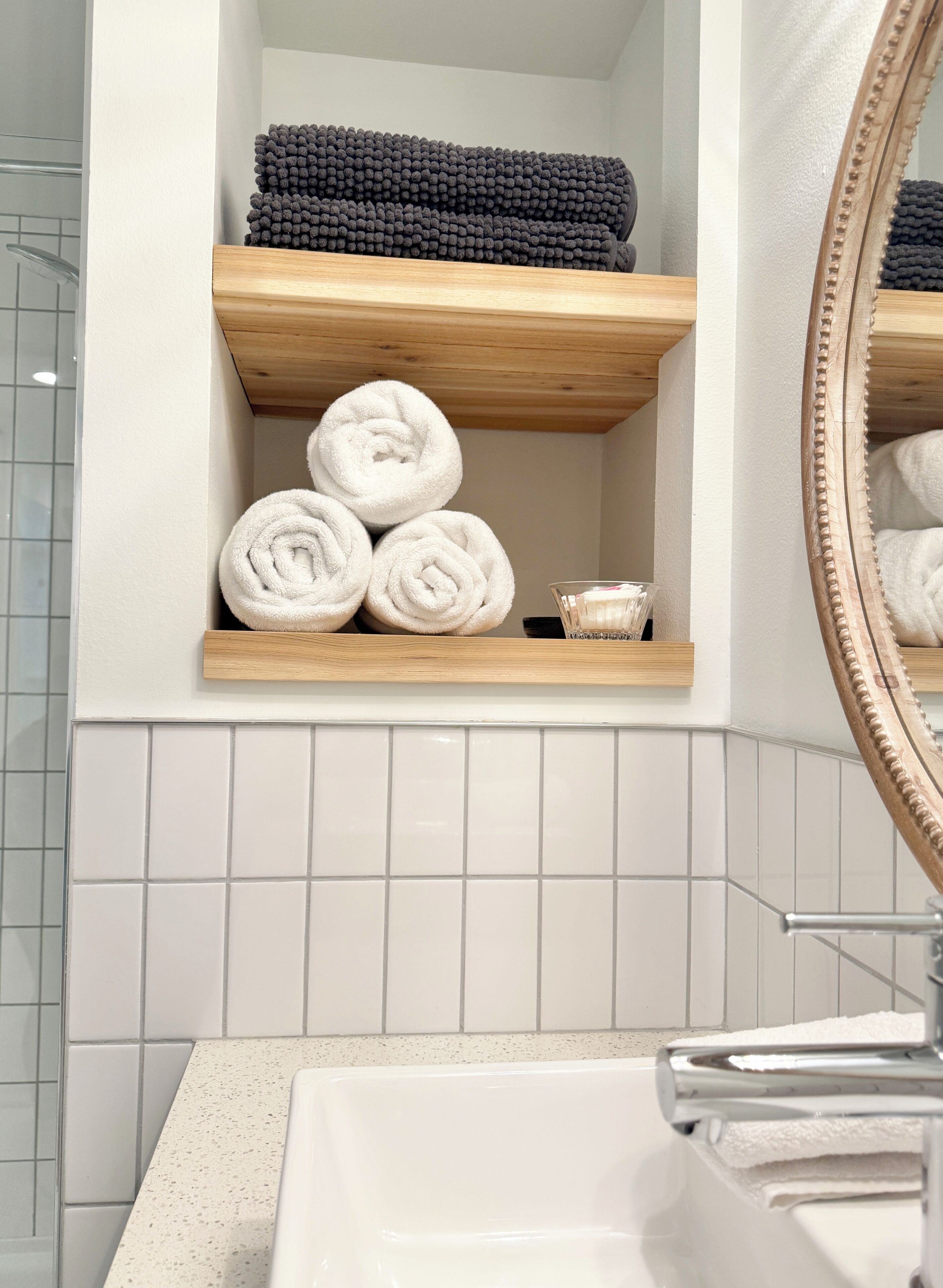 A bathroom with a wooden shelf holding rolled towels and a glass bowl with a bar of soap.