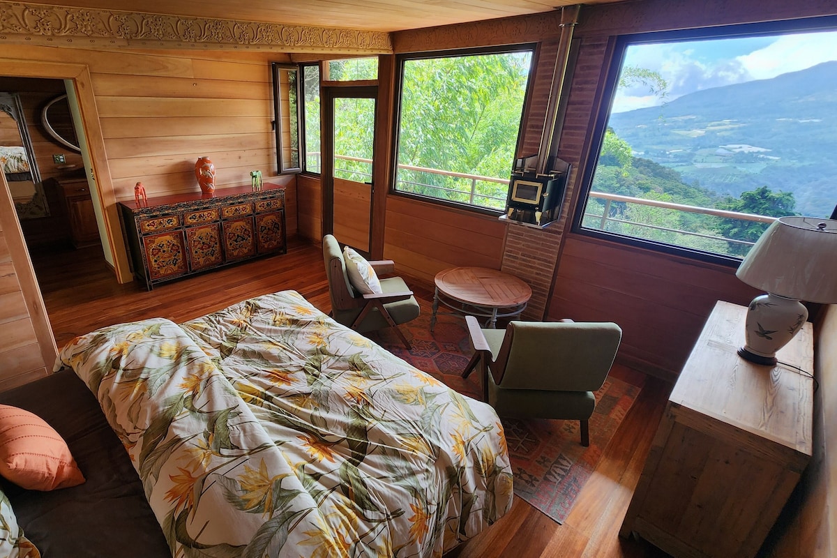 Una habitación tipo cabaña de madera con una cama, una silla, una mesita, un aparador y un ventanal grande que ofrece vistas a la vegetación y montañas.