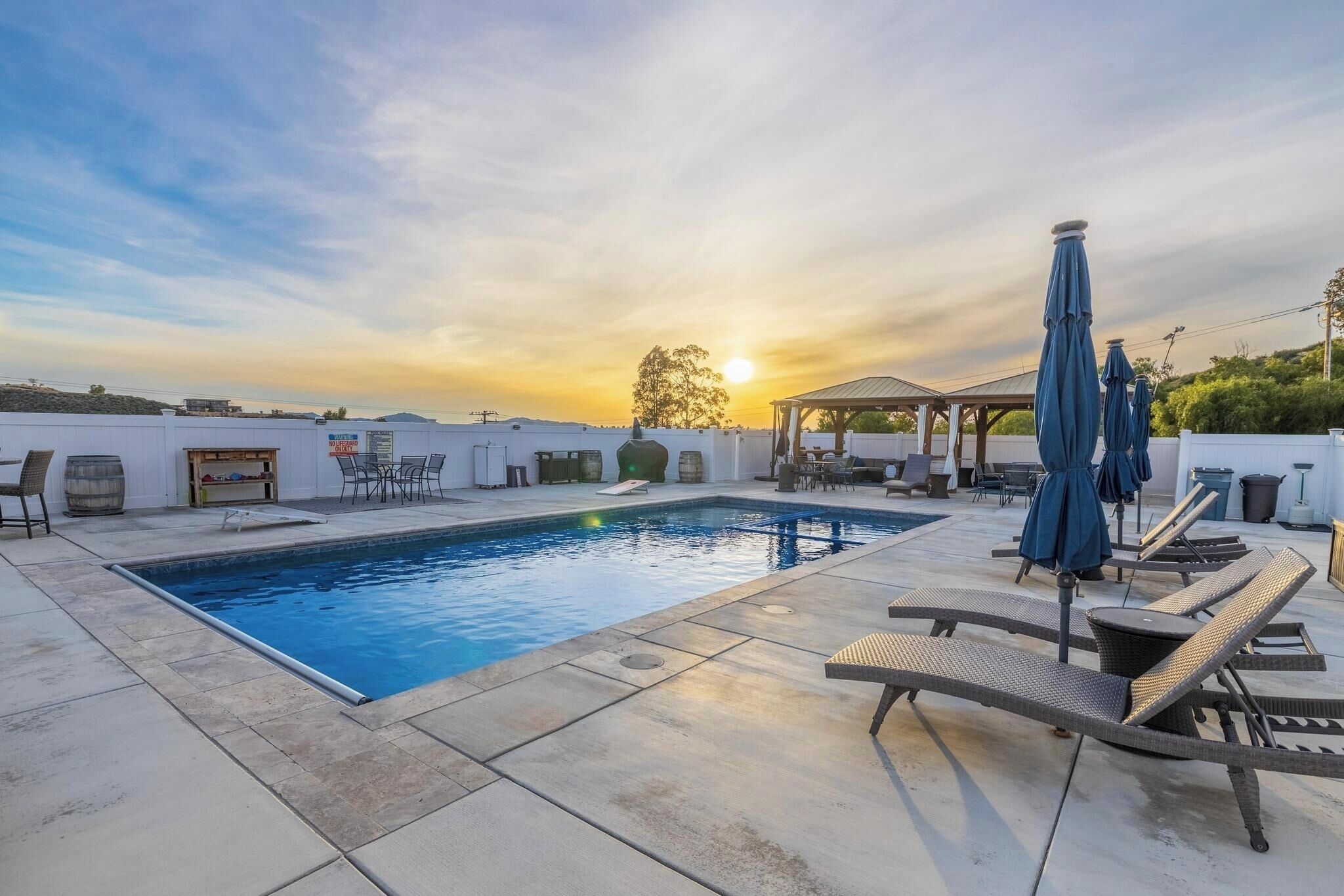 Piscina com cadeiras de descanso e guarda-sóis, cercada por um terraço com móveis de exterior e um gazebo.