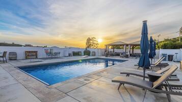 Piscina com cadeiras de descanso e guarda-sóis, cercada por um terraço com móveis de exterior e um gazebo.