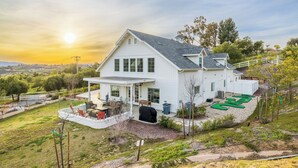 Une maison blanche à deux étages, dotée d’un patio couvert, d’un garage et d’une cour arrière aménagée avec un espace vert pour pratiquer le putting-green.