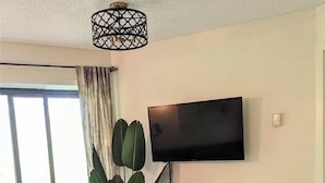 A living room with a flat-screen TV mounted on the wall, a brown chair with a patterned cushion, a wooden coffee table, and a large plant. - Chic ski-in ski-out slopeside 1 bedroom condo at Holiday Valley resort sleeps 4. (Great Valley)