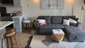 A modern living room with a grey sofa, wooden chairs, a coffee table, and a kitchen area with a stove and cabinets. - Chic ski-in ski-out slopeside 1 bedroom condo at Holiday Valley resort sleeps 4. (Great Valley)