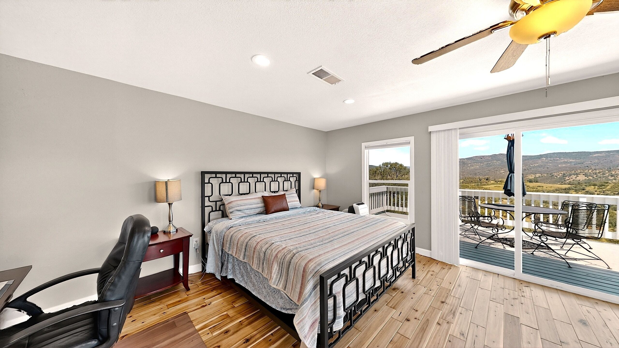 A bedroom with a bed, bedside tables, a chair, a ceiling fan, and a balcony with a view.