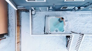 Een hottub in een besneeuwde omgeving, met een houten terras en een blauw gebouw op de achtergrond.