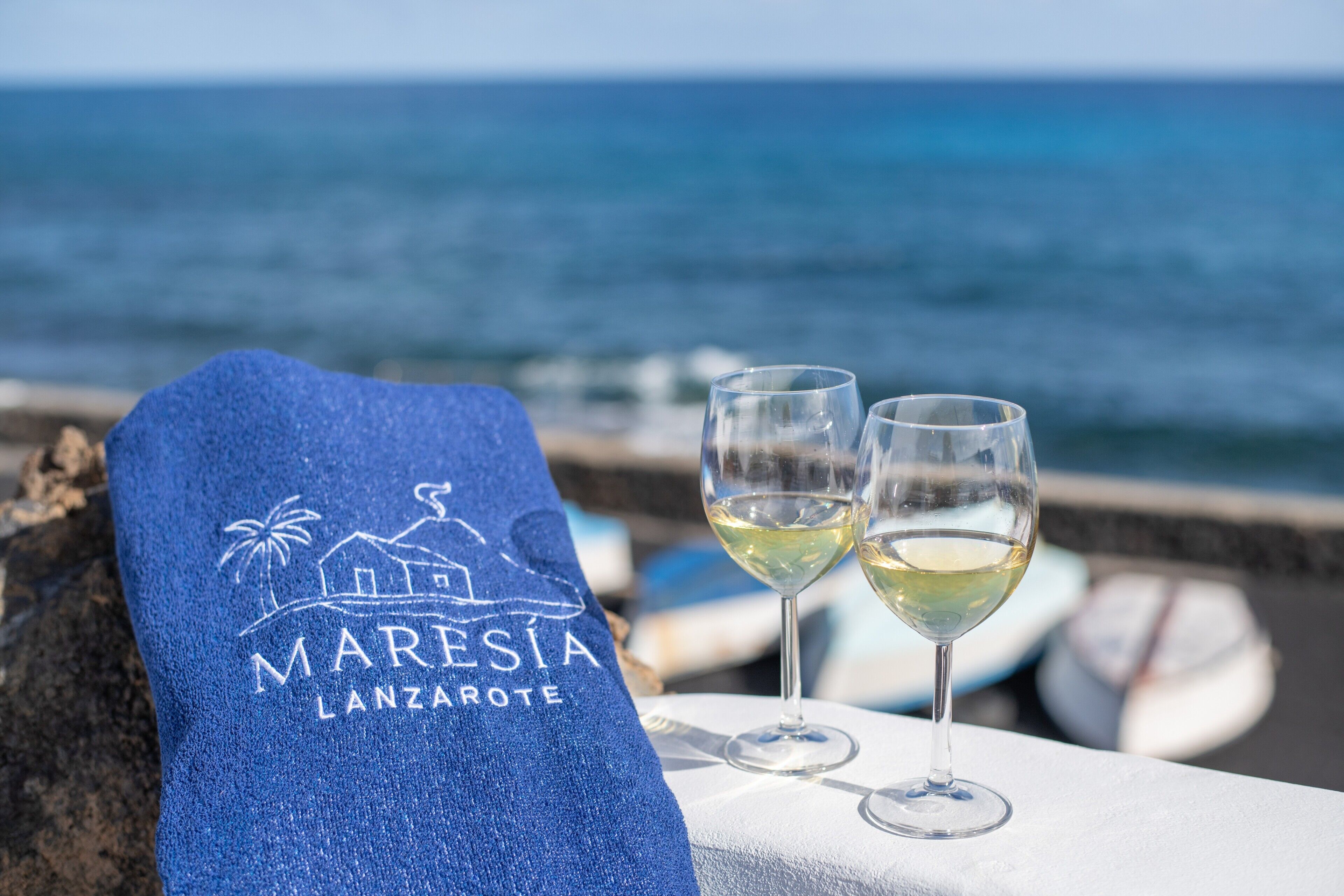 Una toalla con el texto 'MARESIA LANZAROTE', dos vasos de vino blanco y vista al mar.