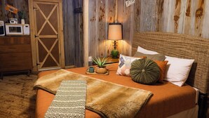 A cozy bedroom with a wooden door, a bed with a woven headboard, a lamp, and a small table with a plant.