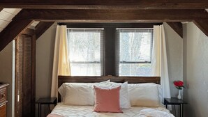 Ein Schlafzimmer mit Holzbalkendecke, einem Bett mit weißer und pinkfarbener Bettwäsche, einem Nachttisch mit Blumen in einer Vase und einem Fenster mit Jalousien.