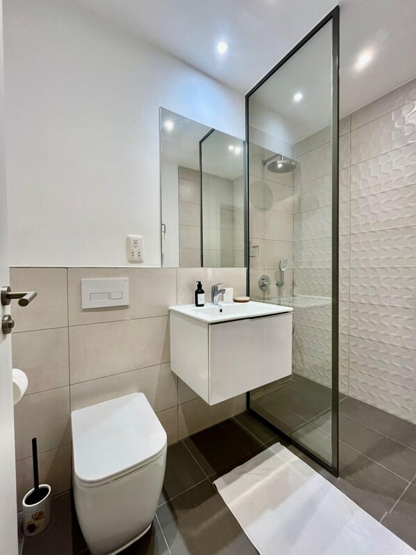 A modern bathroom with a glass shower enclosure, a white sink, and a toilet. - Bantree Luxury Apartments by Cozy (Accra)