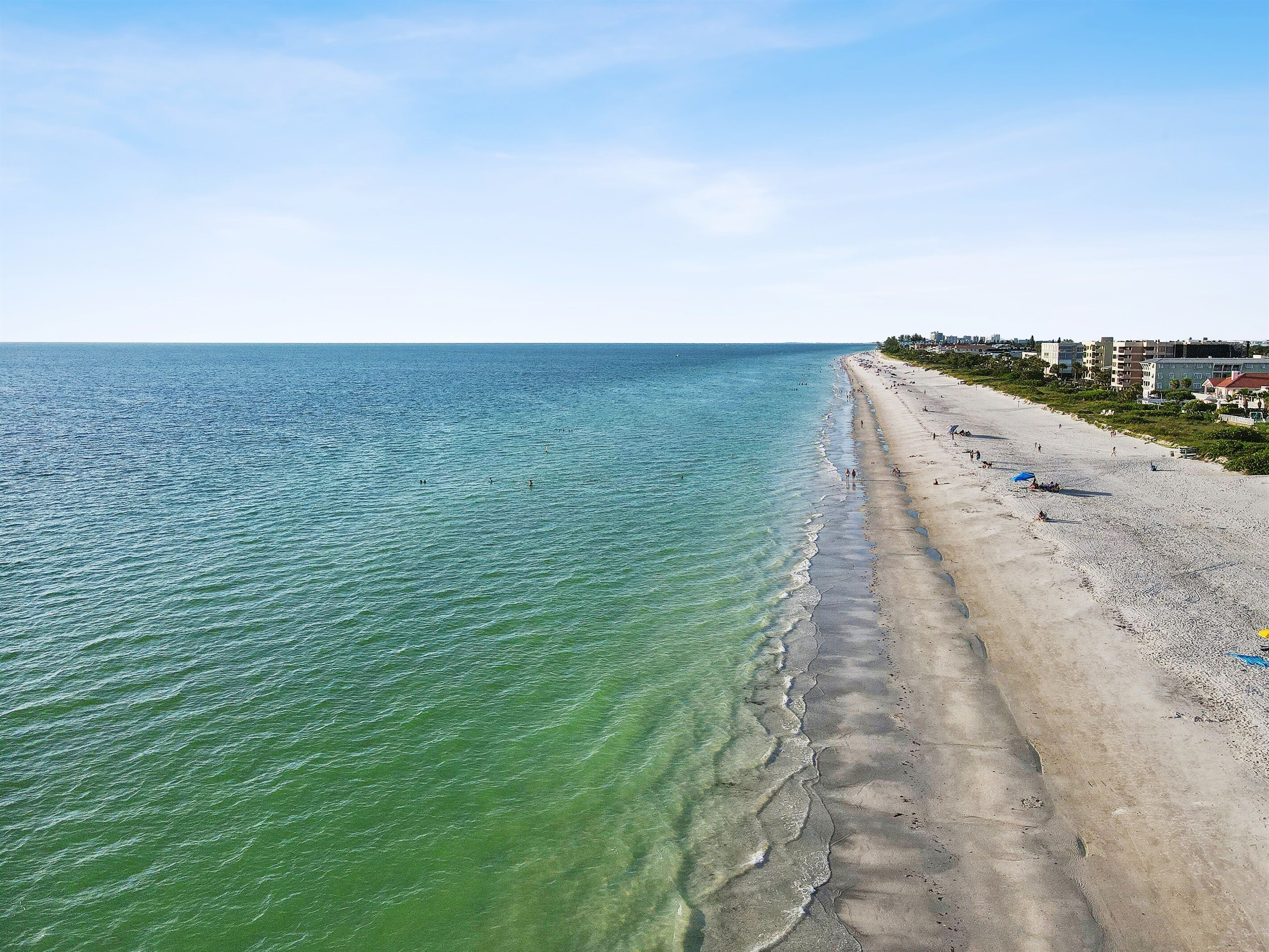 A beach with clear water, a sandy shore, and buildings in the background.