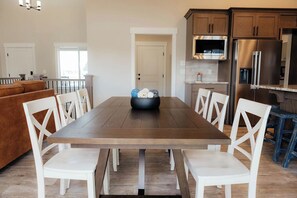 A dining area with a wooden table and white chairs, a kitchen with wooden cabinets and stainless steel appliances, and a view of the outdoors through a door.