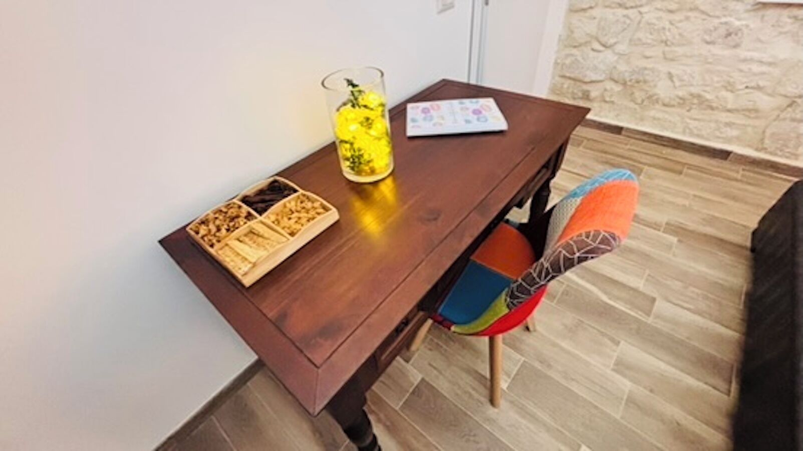 A wooden table with a glass container and a board game, a colorful chair, and a stone wall in the background.