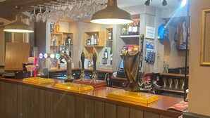 Una zona de bar con paneles de madera, una barra con varias llaves y vasos, y estantes llenos de botellas y aperitivos.