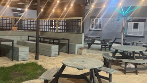 Un espacio al aire libre con mesas de picnic y bancos, rodeado por un muro bajo y un edificio con ventanas.