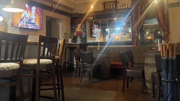 Zona de bar con sillas de madera, un televisor mostrando una escena de fuego y un letrero navideño.