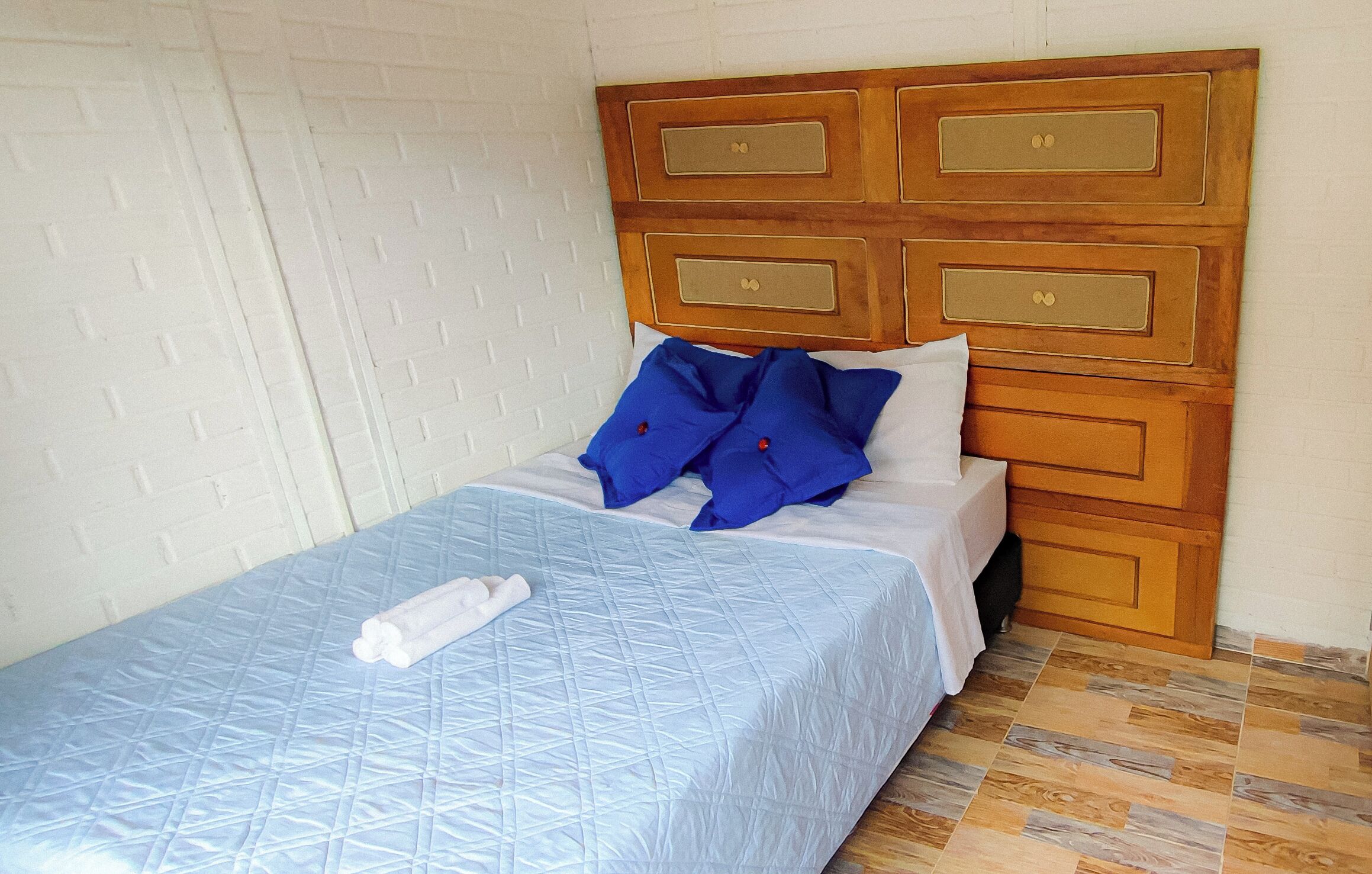 Una cama bien hecha con colcha azul, un cabecero de madera con cajones y una pared con diseño de ladrillos blancos.