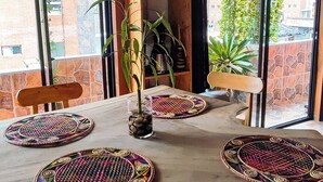 A wooden dining table with woven placemats, surrounded by wooden chairs, in a room with a stone wall and large windows.